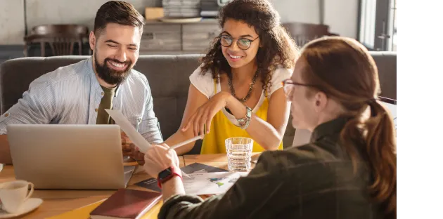 Duas mulheres e um homem sentados ao redor de uma mesa em reunião com um escritório ao fundo.