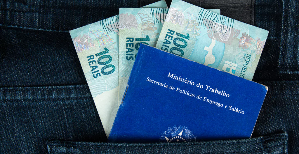 Carteira de Trabalho e notas de 100 reais no bolso de uma calça jeans, representando remuneração e seguro-desemprego.