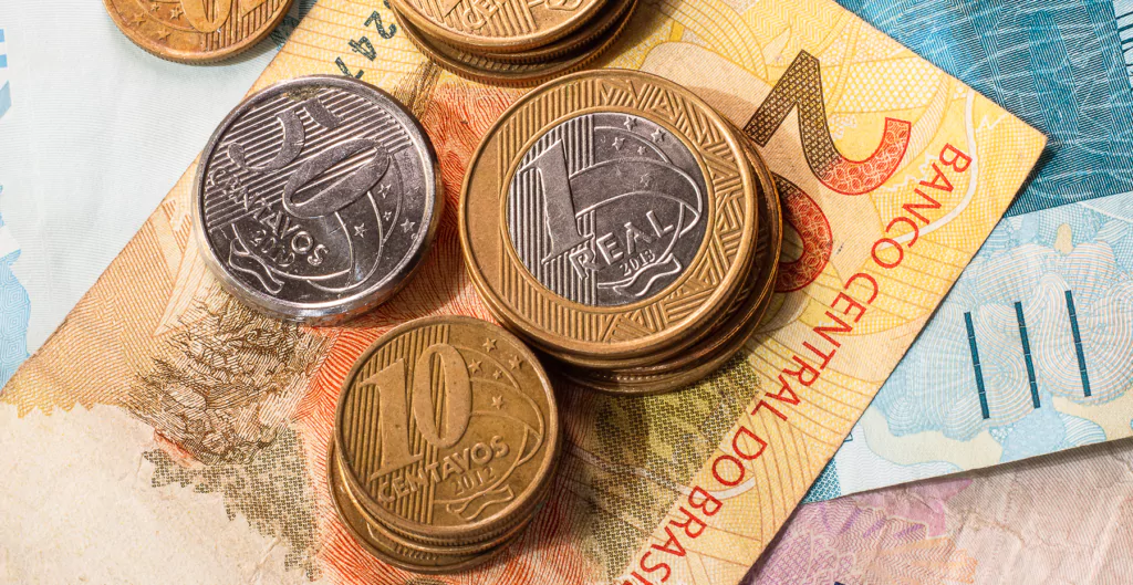 Moedas de 1 real e 10 centavos sobre notas de reais, sugerindo temas de economia e retomada do seguro-desemprego.