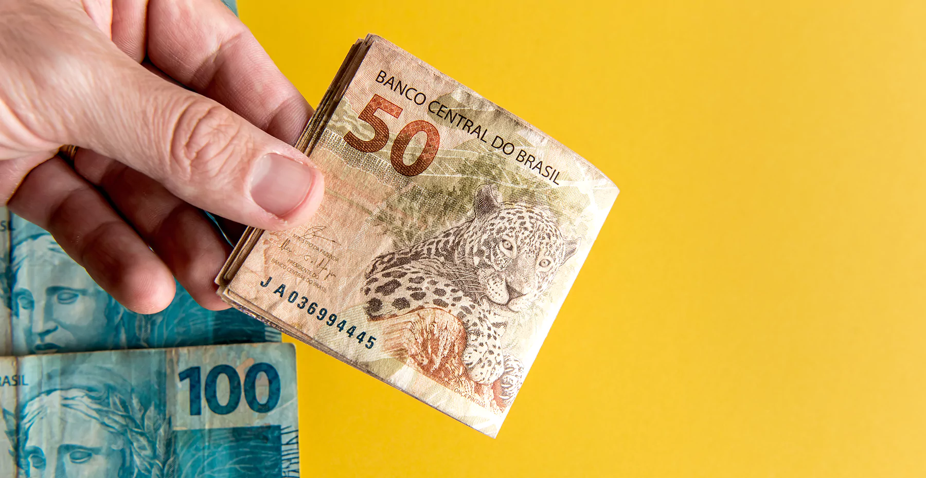 Mão segurando cédulas de 50 reais brasileiras sobre fundo amarelo, simbolizando dinheiro e economia.